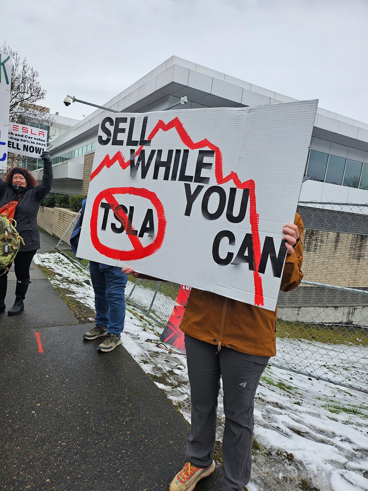 A sign that reads "SELL WHILE YOU CAN" over a red downward trending stock price chart line, along with Tesla's stock ticker TSLA with a circle and a line through it, being held by a protester. There is very light snow on the ground, and other protesters are visible