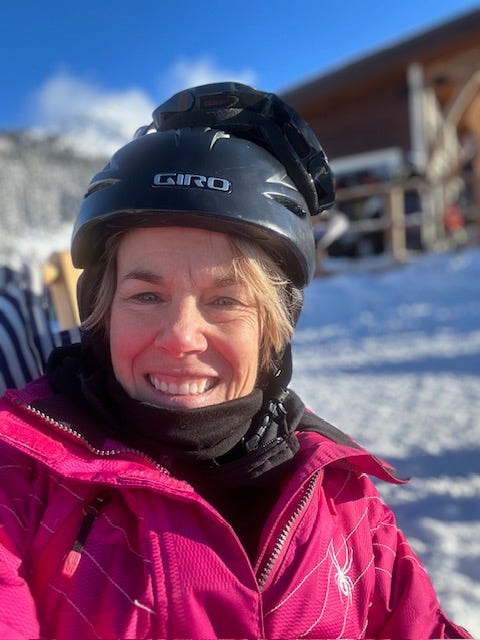 A smiling woman sitting at a ski hill, she is wearing a black helmet and pink ski coat.