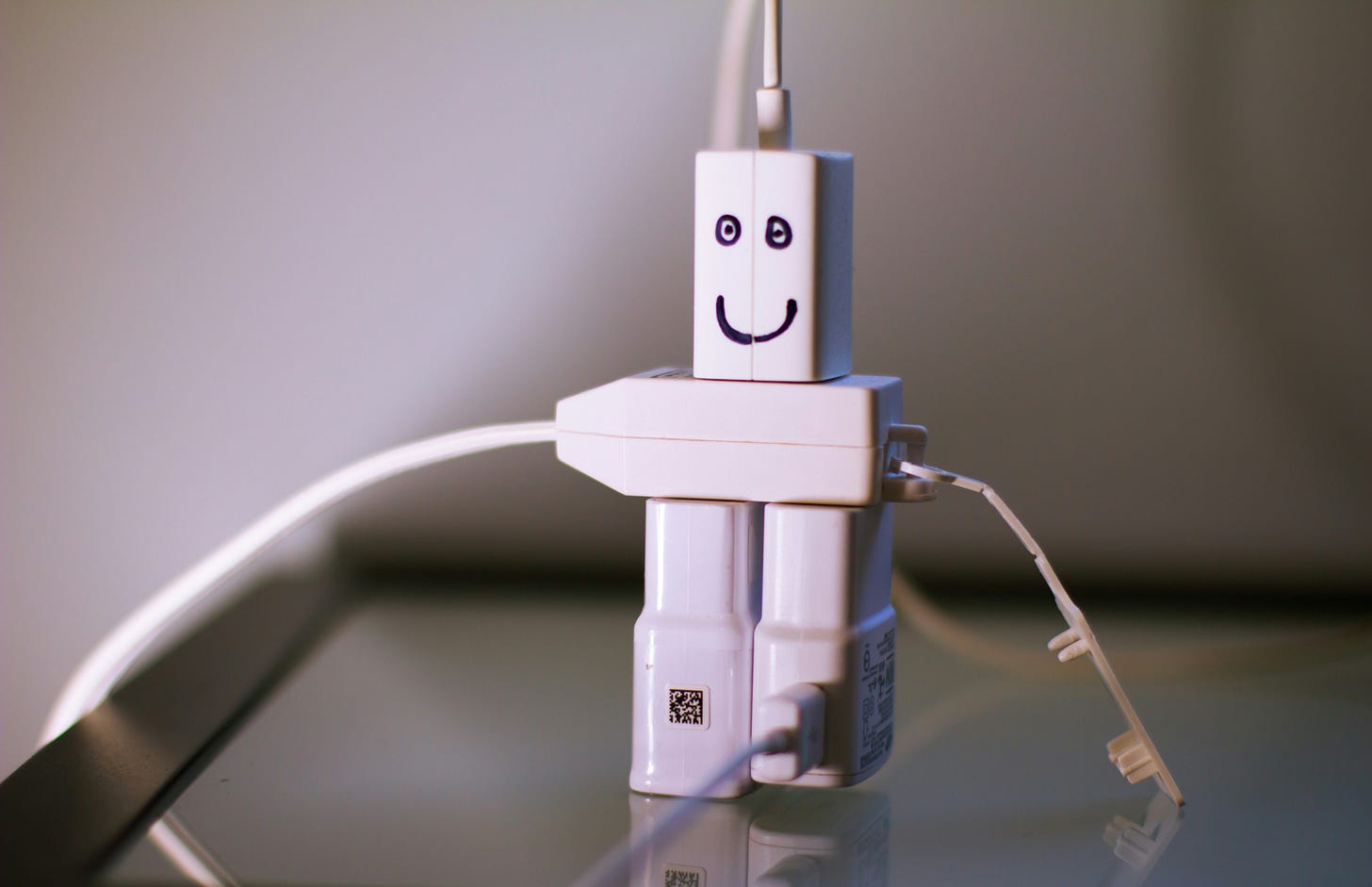 A photo of several white chargers and extension cords plugged together in the shape of a person. There is a smily face drawn in marker on the "head" of the figure.