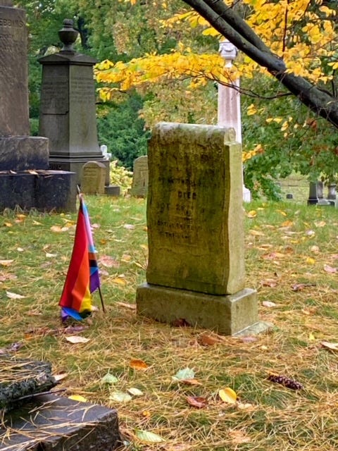 A badly-weather gravestone in a well-populated cemetery by a tree with golden leaves. To left of it, a Progress Flag has been planted in the ground.