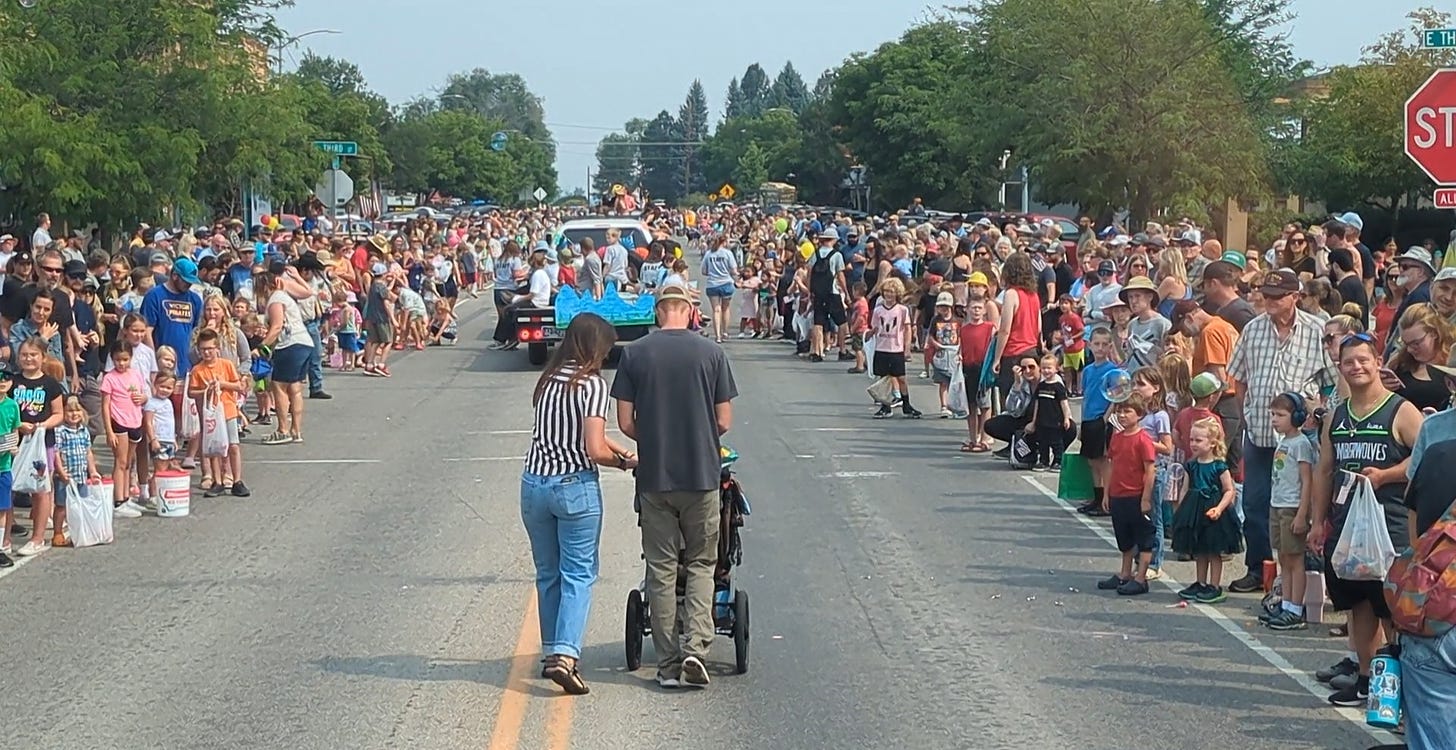 a crowd of people gathered along both sides of a street with a float in the distance and a young couple pushing a stroller in the foreground