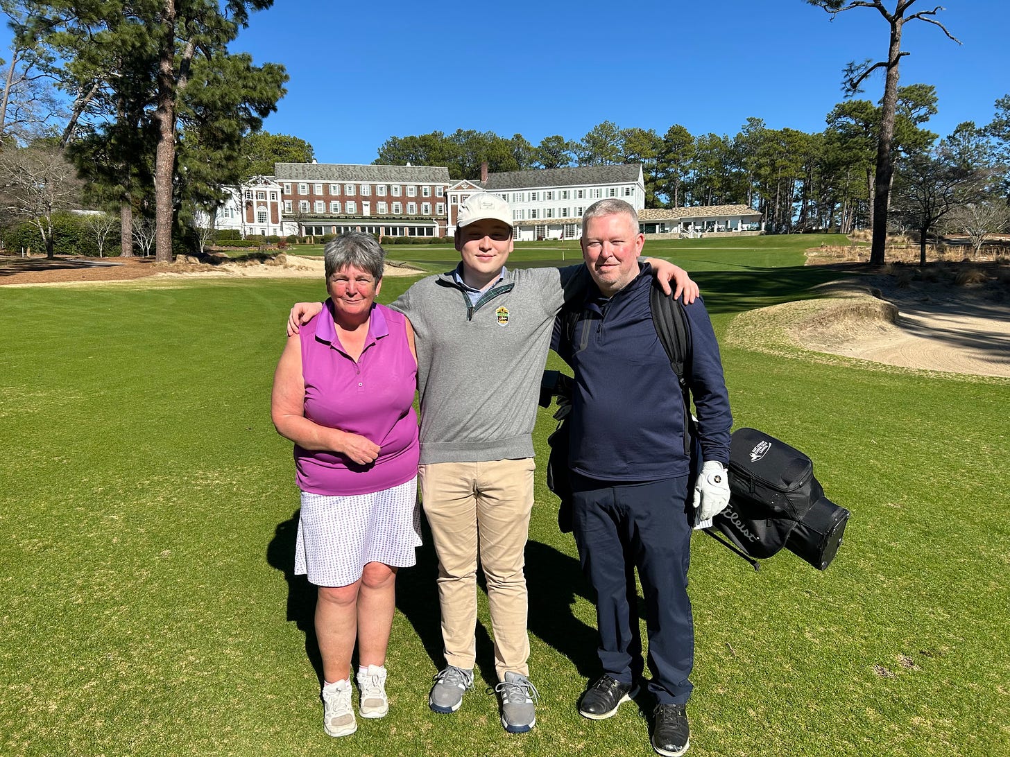 Shona, Jake, and Steve stand just short of the 18th green at the Mid Pines Golf Club. In the background, the Mid Pines Lodge imposes.