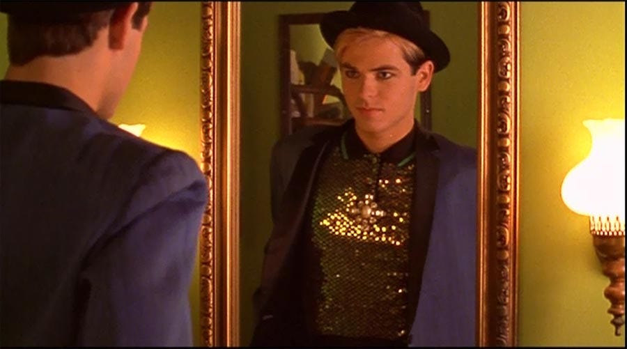 A white teen guy looks in a gold-framed mirror. He has dyed blonde hair and black eye makeup, with a rimmed black hat, purple blazer, and gold sequined shirt. He is looking at himself with self affirmation before leaving the house for a night out.