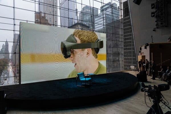 Mark Zuckerberg is on a large screen on a stage demonstrating virtual reality technology. The large window behind the screen shows a street in Manhattan.