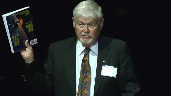 A man with white hair wearing a suit with a name tag. He holds up a book 