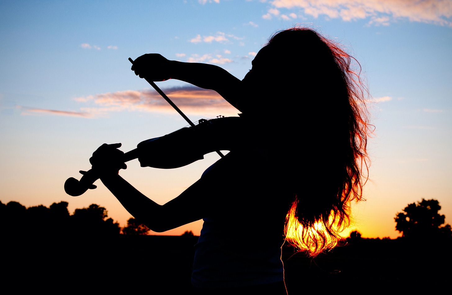 Long-haired young woman playing violin, silhouetted against the sunset in nature