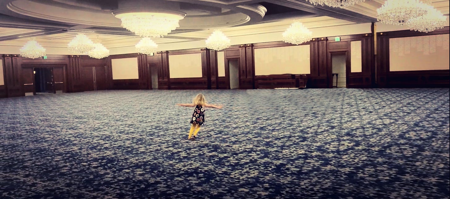 blonde child in donut dress and yellow pants twirls in a large, empty ballroom