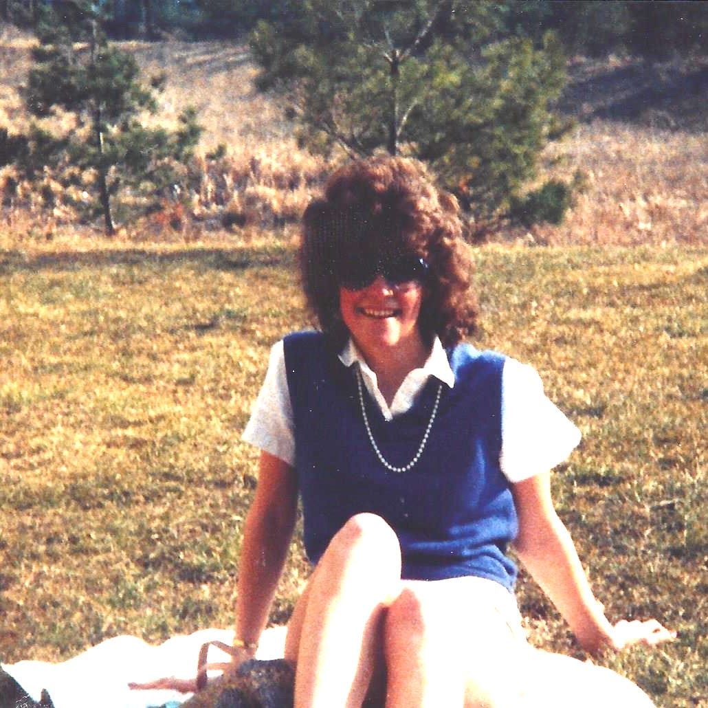 Young college-aged girl with big hair and hue sunglasses is lounging on a lawn. It must be the 80s