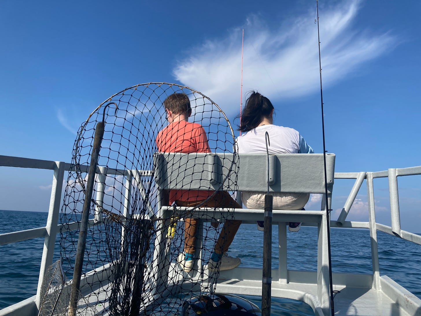 A picture of the back of a boat where two people (a man in an orange shirt and yellow pants, and a young woman with black hair, a white shirt and a pair of shorts) sit facing away from the camera. In the foreground, a large fishing net waits to be used.