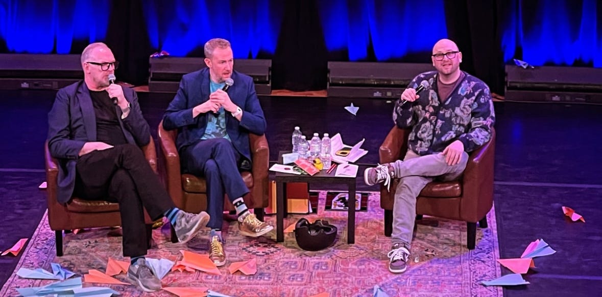 Greg Davies, Alex Horne, and me sitting onstage at Town Hall in NYC. We are surrounded by pink and blue paper airplanes strewn about the stage that have been thrown by audience members.