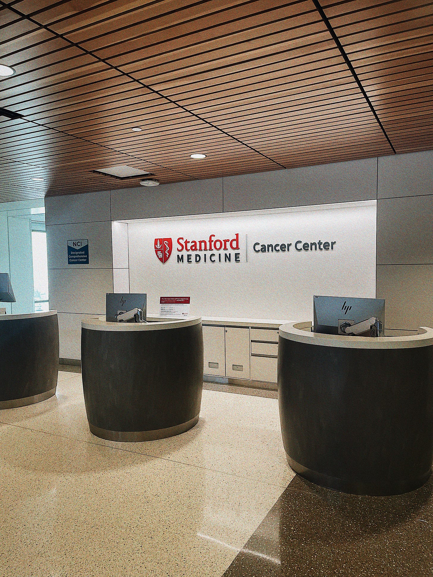  Lobby of the Stanford Medicine Cancer Center with three curved reception desks, a wooden slat ceiling, and polished flooring.