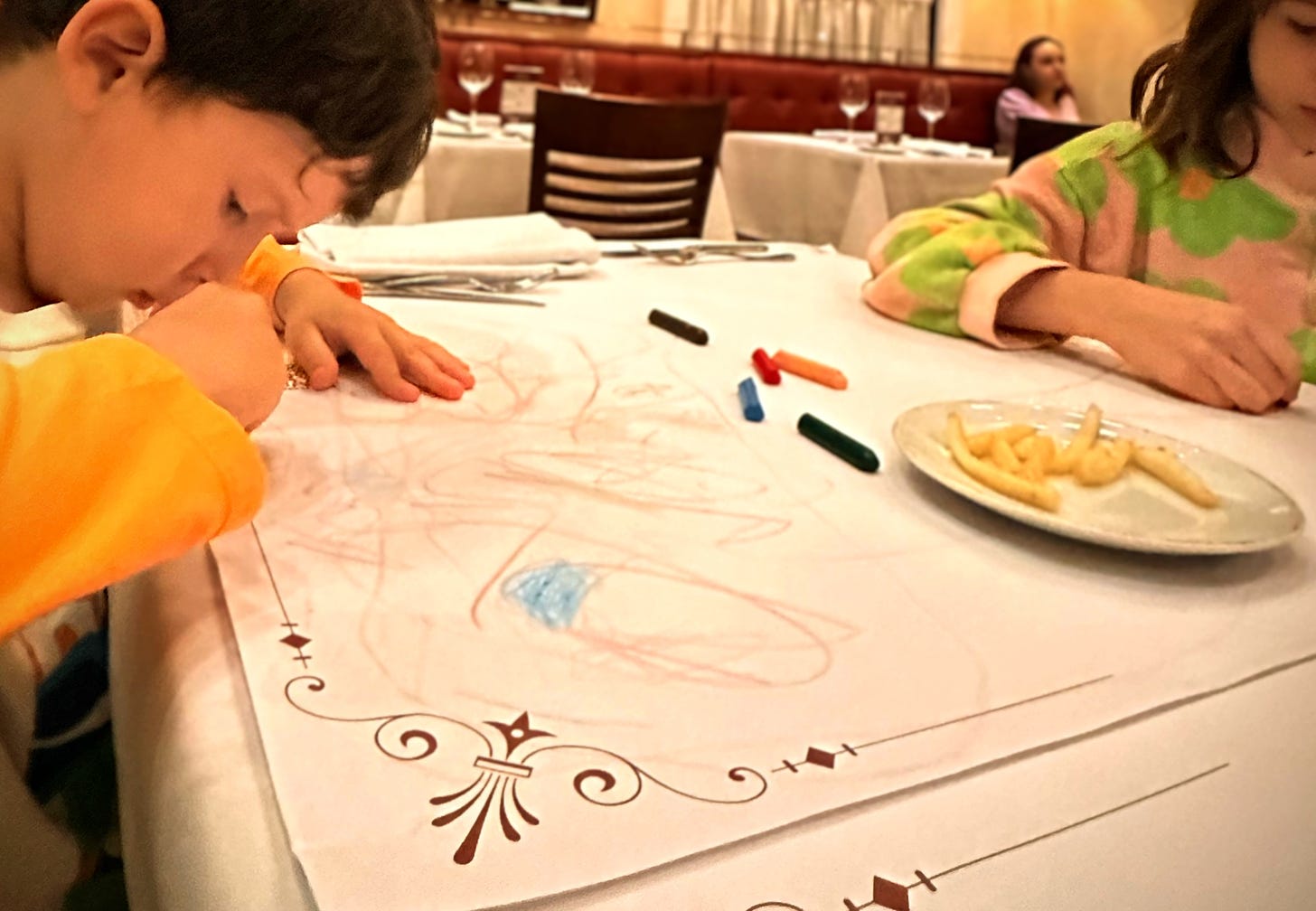 duas crianças desenhando com giz de cera na mesa de um restaurante e uma porção de batata frita