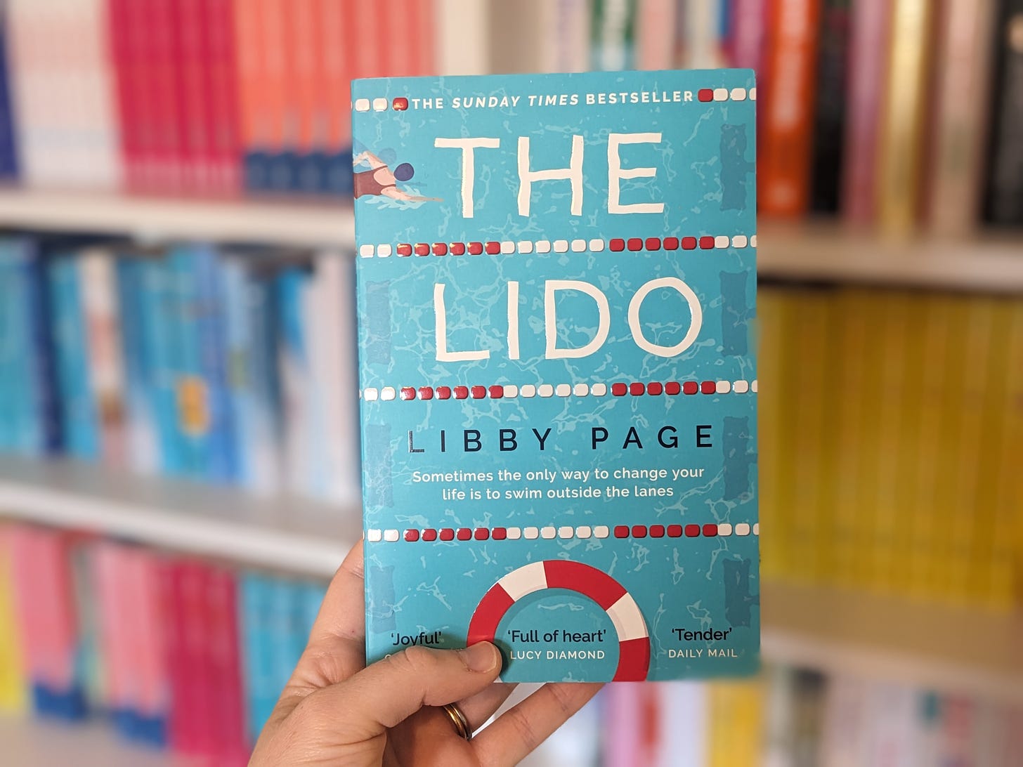 The cover to The Lido by Libby Page shows a birds eye view of a pool with a swimmer, including ropes and a life ring.
