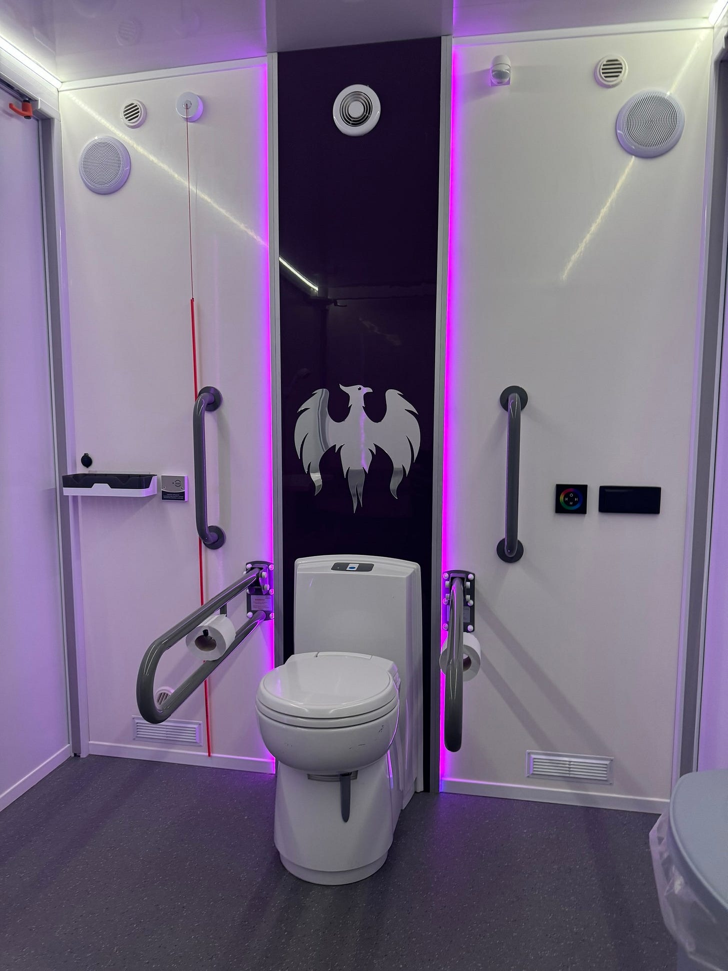 Inside the Mobiloo bathroom.  Lit with blue/pink LED lighting is a fully accessible toilet and sink.  The backsplash for the toilet has a white phoenix motif.  Floors are clear, clean and grey.