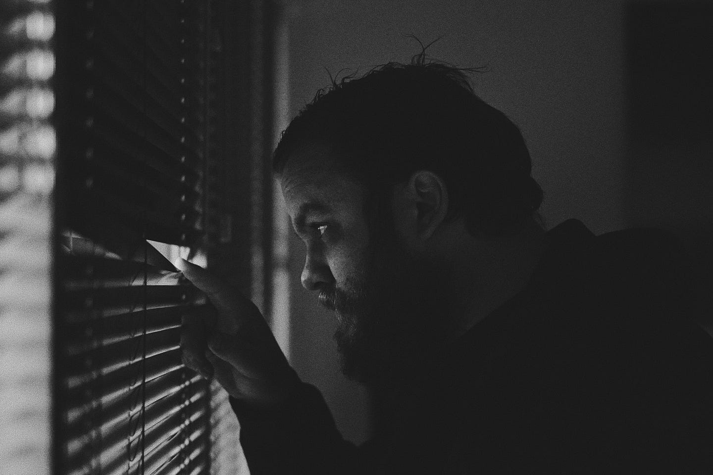 Man in darkened room peeking through blinds toward outside