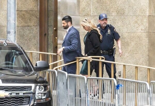 A blond woman leaving a courthouse and walking toward a black Chevrolet SUV. A guard stands behind her and a man wearing a suit is walking in front of her. 