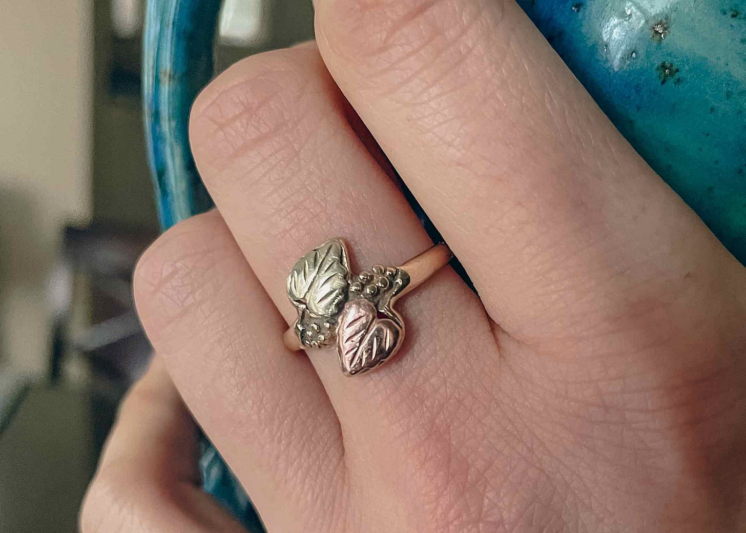 A gold ring with two leaves poined away from eachother, one yellow gold, the other rose gold. A woman wears the rings on her middle finger. Her hand holds a blue coffee mug.