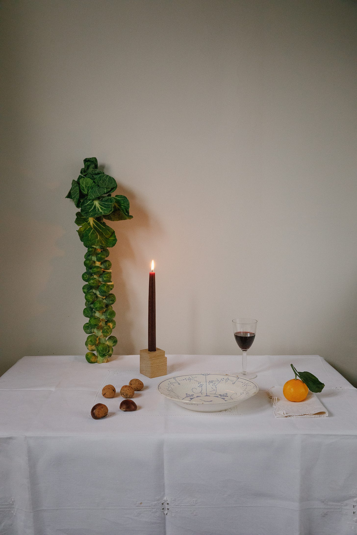 a dinner table scene with a tablecloth, a stick of sprouts, a lit candle in a wooden candle holder, a glass of red wine, a bowl, some cracked walnuts and an orange.