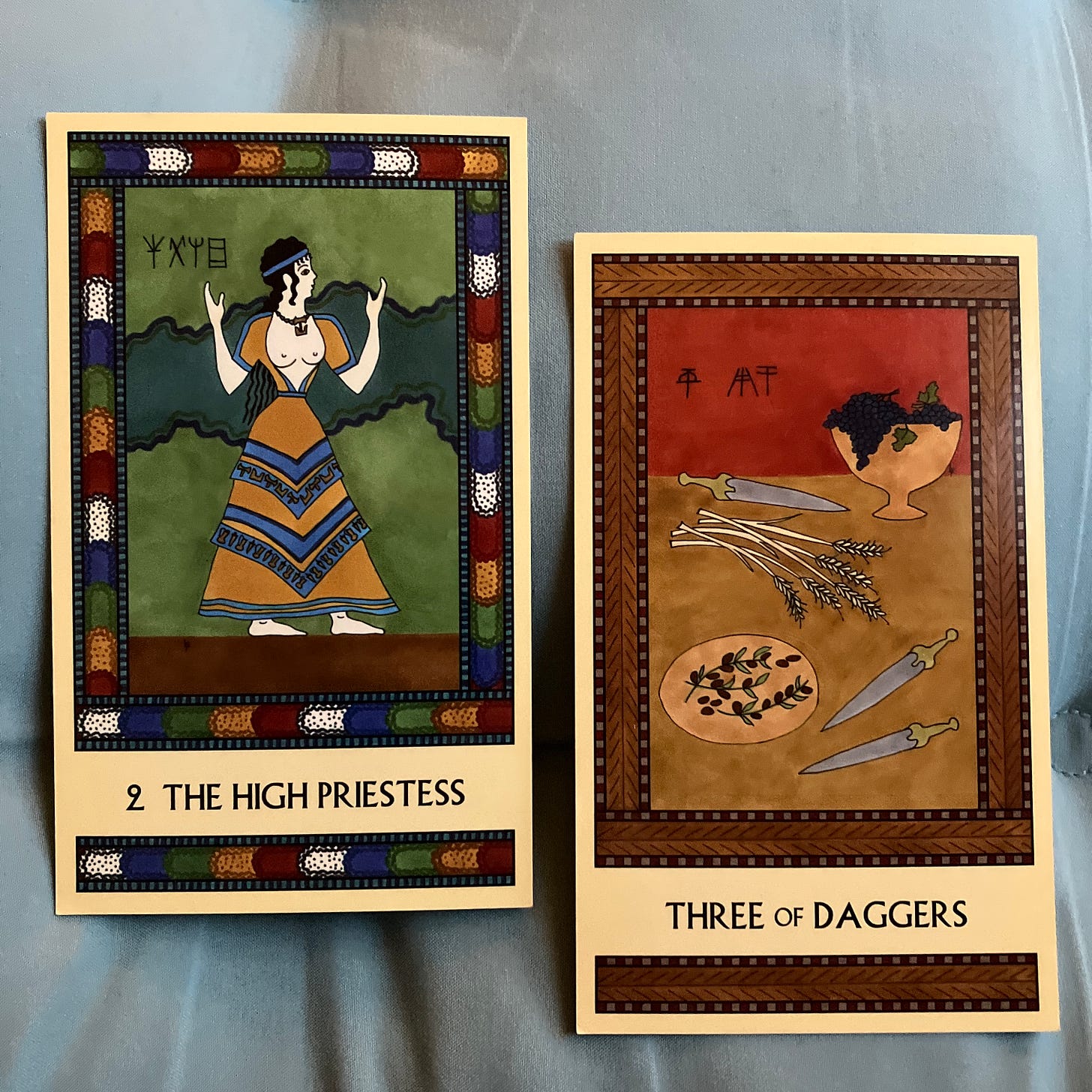 Two Minoan Tarot cards side by side. The High Priestess shows a white-skinned woman with long black hair, wearing a Minoan-style top and skirt in shades of yellow and blue. She faces right with her arms raised in a sacred gesture. The Three of Daggers shows a table laden with a platter of olives, a pile of grain stalks, a footed bowl full of grapes, and three gold-hilted daggers. 