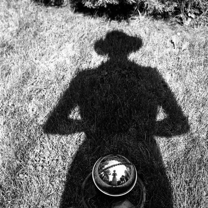 Uma pessoa está em um campo gramado, com sua sombra se estendendo diante dela. O sol está brilhando intensamente e o céu é de um azul brilhante. Em uma das mãos, ela segura uma câmera fotográfica antiga, enquanto na outra segura uma bola metálica que reflete seu corpo bem pequeno, quase indistinguível. A cena é emoldurada por árvores que se erguem contra o horizonte, lançando longas sombras no chão.