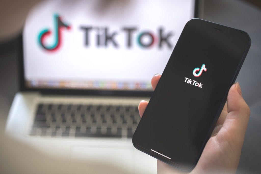 A black iPhone held in a hand with the TikTok logo displayed on the screen in front of a silver macbook open to TikTok on a web browser
