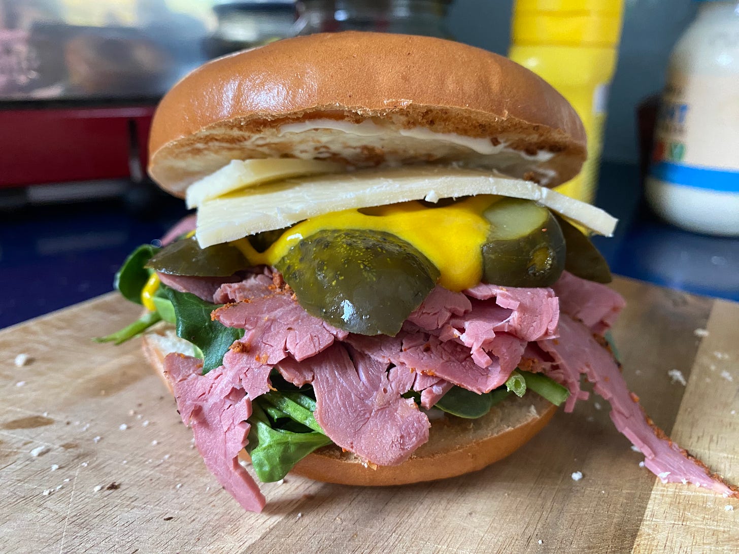 Bagel sandwich filled with pastrami, lettuce leaves, pickles, mustard, and cheese. The bagel is placed on wooden board