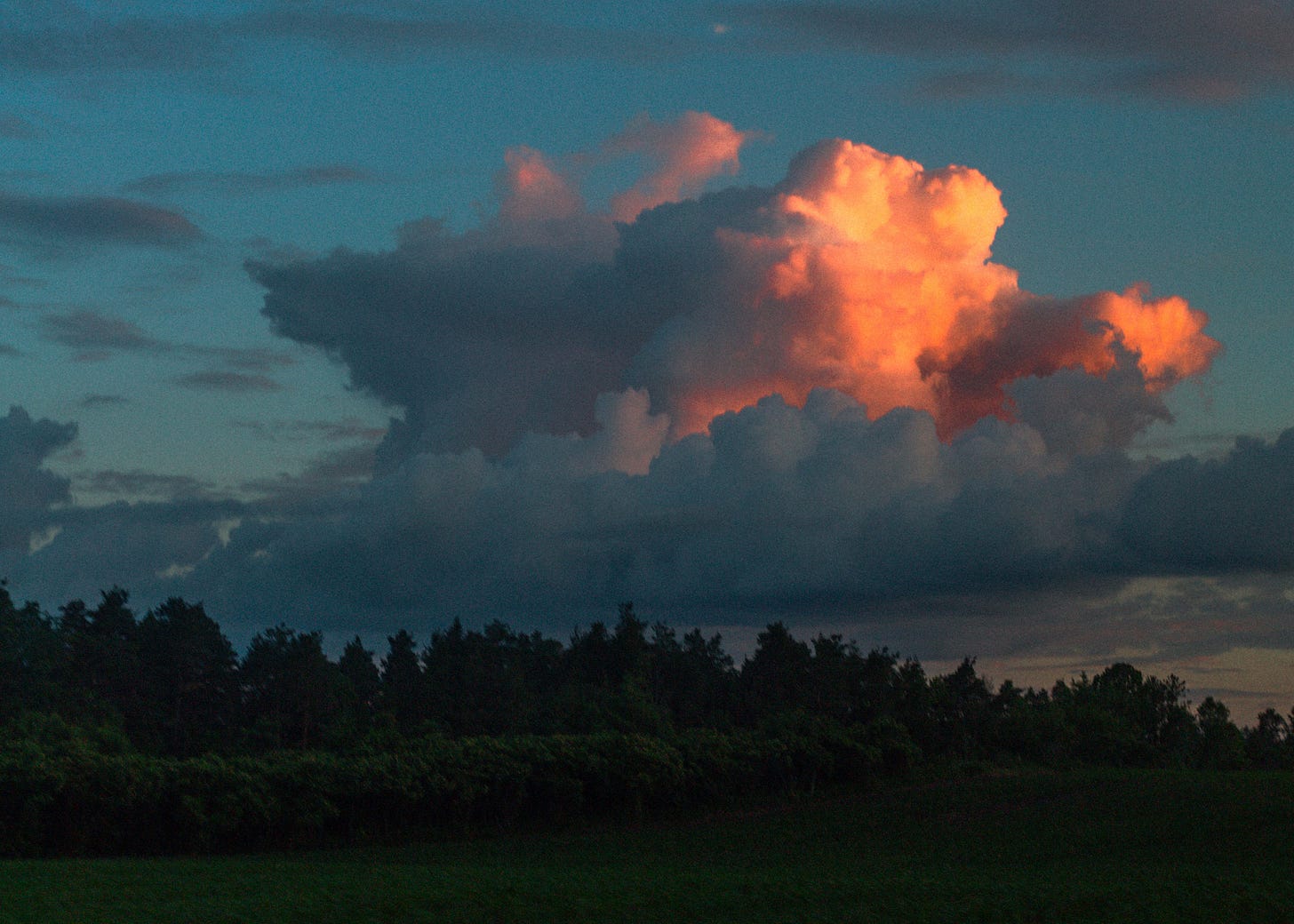 A digital photograph of a landscape. A large cloud floats in the sky, half of it bright orange with the sunset. Below is a deep green horizon of trees. Shot by Rachel, 2023