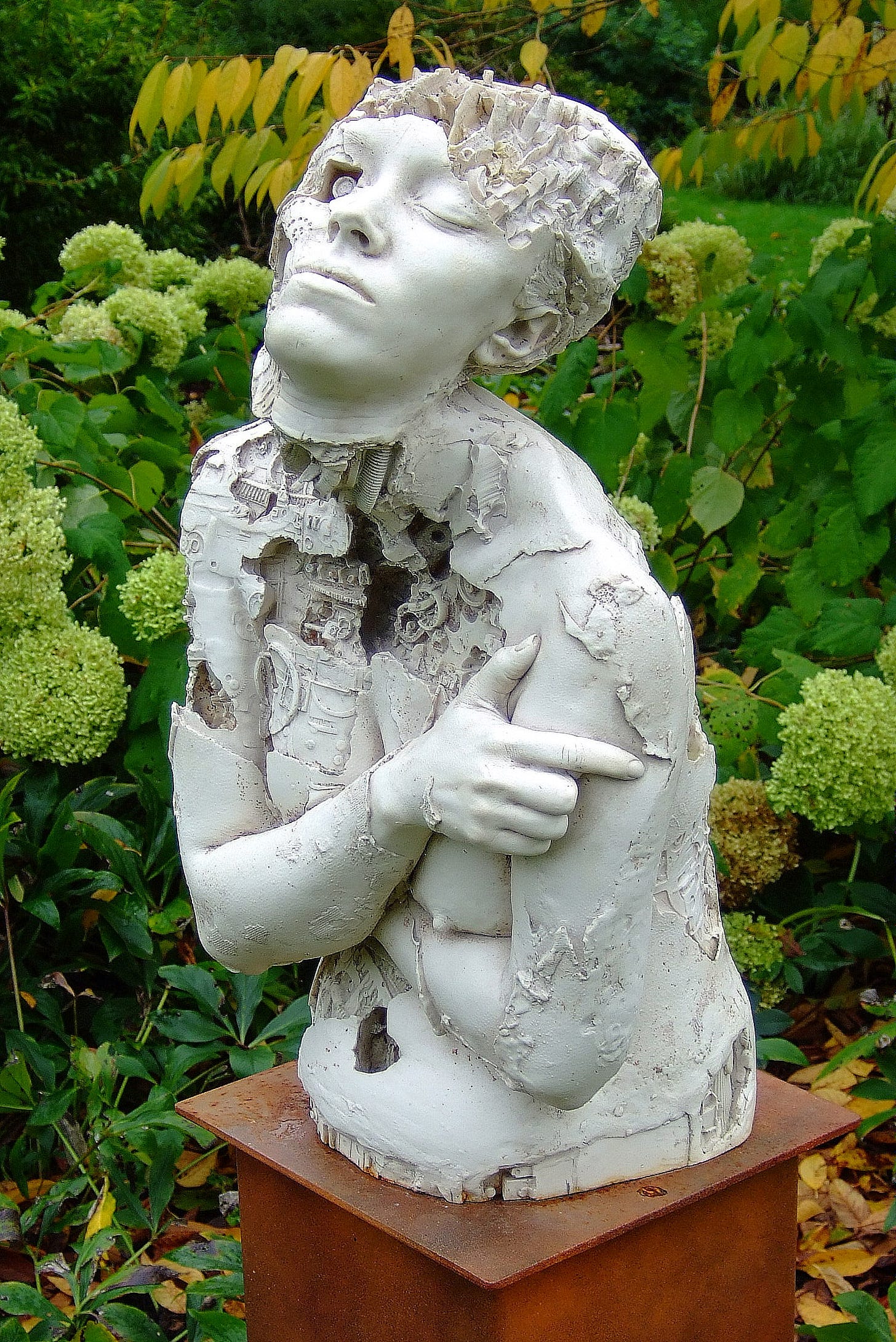 A photograph of a marble resin sculpture of a cyborg mounted on a simple wooden plinth stood in a garden setting. The concepts of time and discovery are intended to be portrayed in this sculptural work. Machine made objects creating mechanical textures have been fused with a human form cast from life. The skin has eroded in areas to reveal mechanical systems analogous to the internal workings of the body.