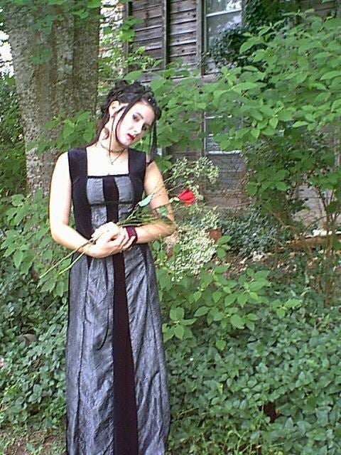 Middle school Lyric, in a silver and black prom-dress, with their face painted white, dark eyes, and bright red lipstick, they pout, holding a red rose, with a maroon band on their wrist.