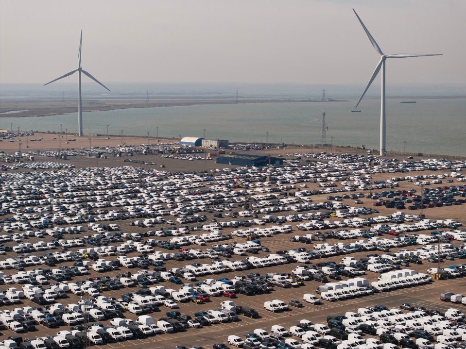 Aerial view of a large car and van storage lot at Peel Ports Ltd. in Sheerness, England, with wind turbines in the background.