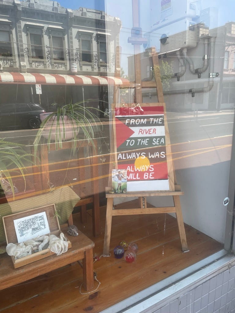 photo of the storefront window of theyswarm, with artwork with the Palestinian and australian Aborliginal flag and the words FROM THE RIVER TO THE SEA ALWAYS WAS ALWAYS WILL BE