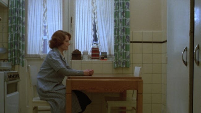 Still from Jeanne Dielman actor Delphine Seyrig sits at  a kitchen table and stares 