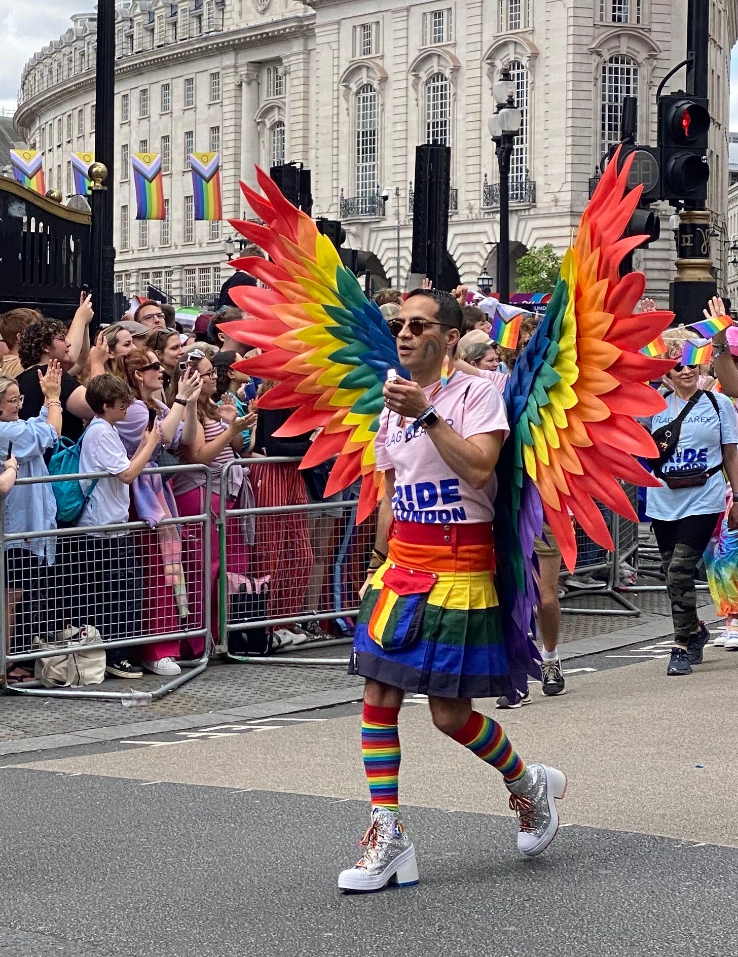 a person in a rainbow skirt and with rainbow wings and rainbow knee-high socks walks down a street near Piccadilly Circus in Sparkly high boots while onlookers standing behind the pedestrian fence look across to the right at other people walking in the parade