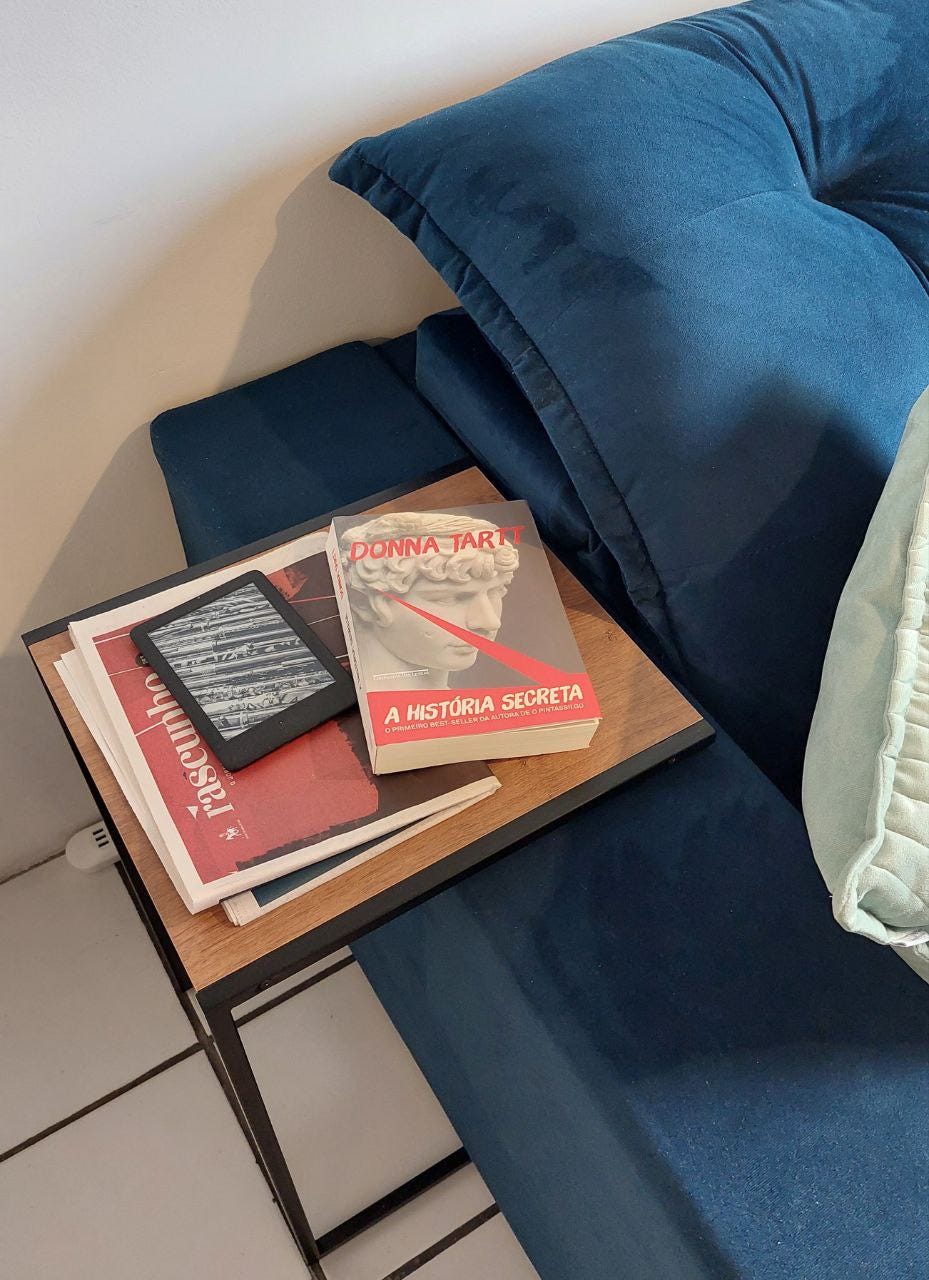 Uma foto da minha sala, mostrando um canto do sofá azul e uma almofada verde água à esquerda. Uma mesinha lateral de tempo de madeira e pés de metal preto. Sobre a mesinha, o livro "A História Secreta", um Kindle preto e uma edição do jornal Rascunho.