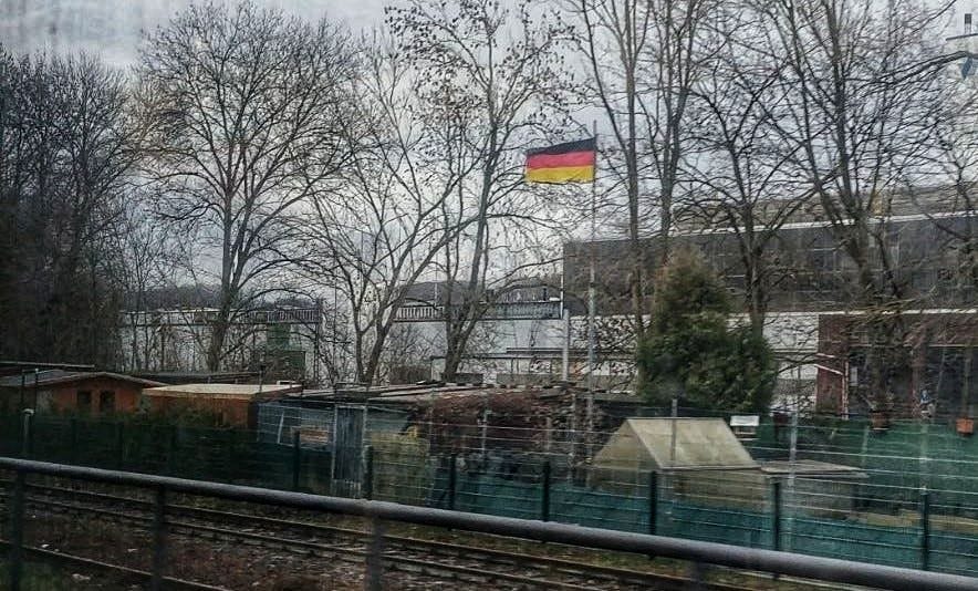 Titelbild: An einer Bahnstrecke weht eine Deutschlandfahne im Wind. Sie steht in einem Schrebergarten neben den Gleisen. Das Foto wurde aus einem Zug heraus gemacht. Der Himmel ist grau, im Hintergrund Fabrikgebäude, es ist Dezember.