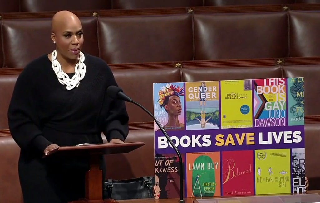 Video Screenshot of Ayanna Pressley speaking in House chamber to introduce her 'Books Save Lives' Act. Next to her is a poster with that slogan, with cover images of banned books including 'All Boys Aren't Blue,' 'Gender Queer,' 'Beloved,' and others.