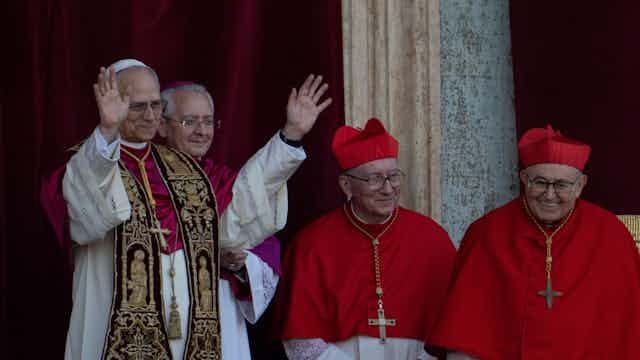 A man in brocaded vestments waves with both his hands while some others dressed in red shoulder capes over white robes smile. A man in brocaded vestments waves with both his hands while some others dressed in red shoulder capes over white robes smile.