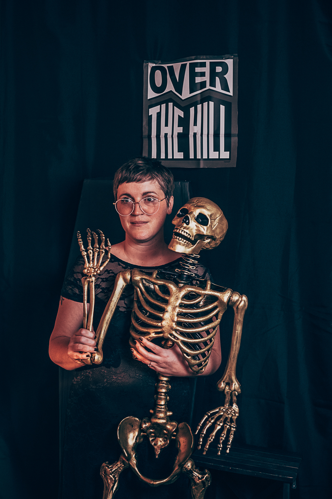 a black background with a sign that says over the hill. a middle aged white person with short hair and round glasses is holding a gold skeleton and making a slightly weird smile at the camera.