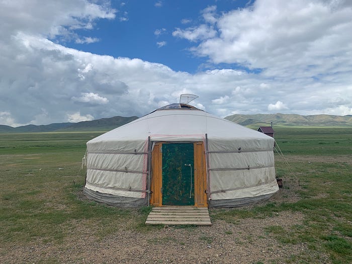 A large round yert style of tent with a door open in the center and a wooden plank platform as steps. It is in a large expanse desert-like plain with hills in the background.