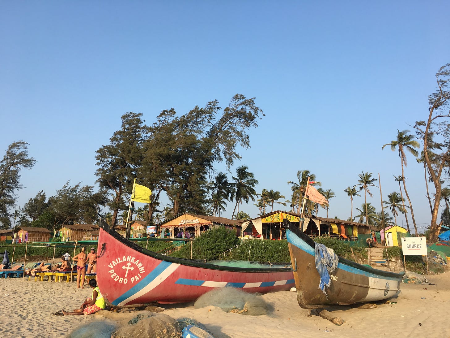 Two fishing boats are docked on the beach. Behind and beside them are beachgoers, palm trees, and several huts. 