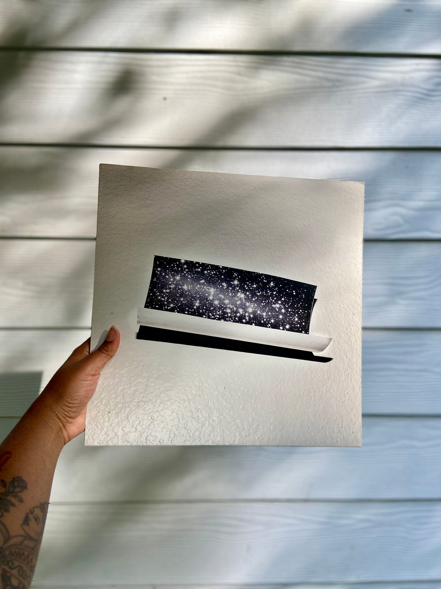 a Black person with a tattooed arm holds a vinyl of Bear in Heaven's fourth album Time is Over One Day Old against a white house. the album has a photo of constellations in the night sky against a textured white wall.