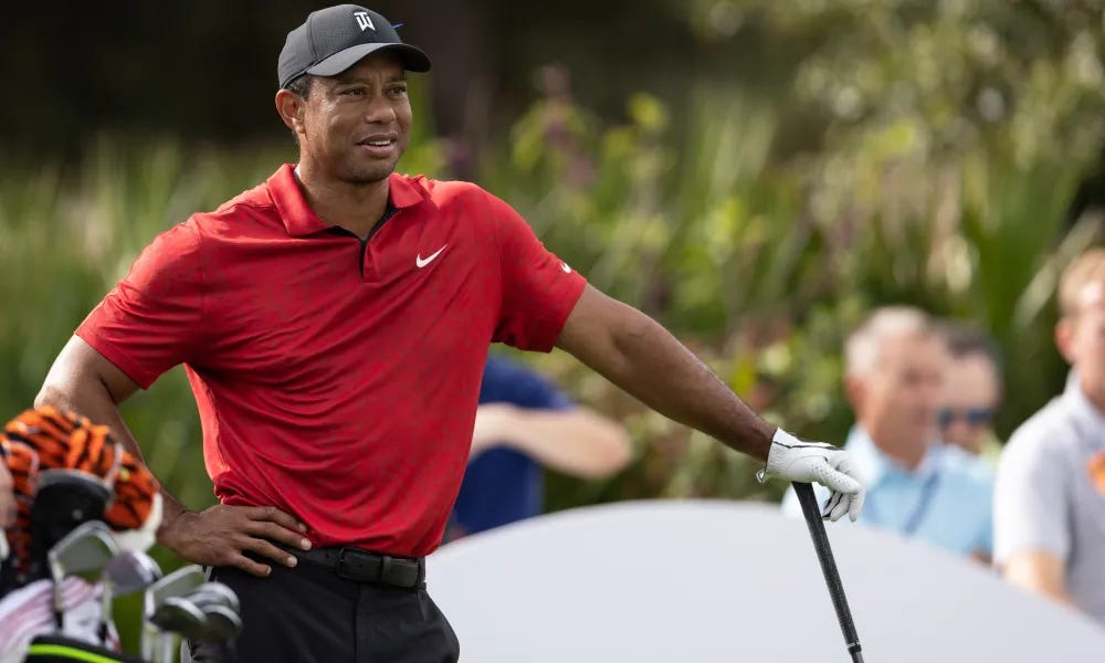 Tiger Woods standing on a golf course, wearing a red Nike polo shirt and black cap, holding a golf club.