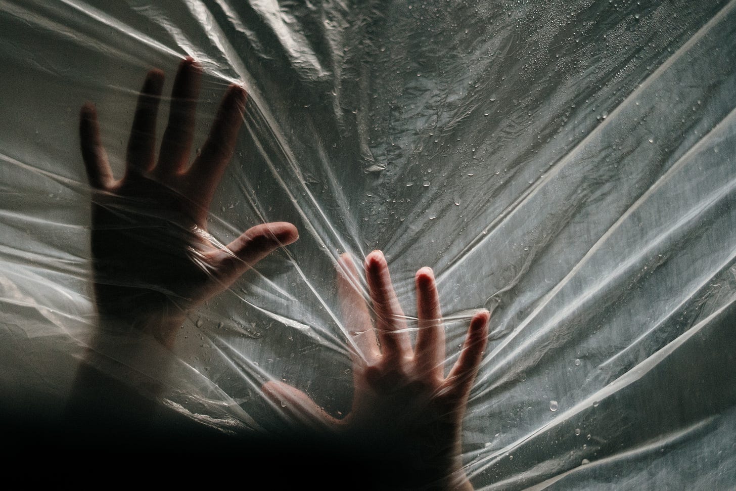 two hands pressing into a sheer, translucent plastic sheet, as if trying to push through to the other side