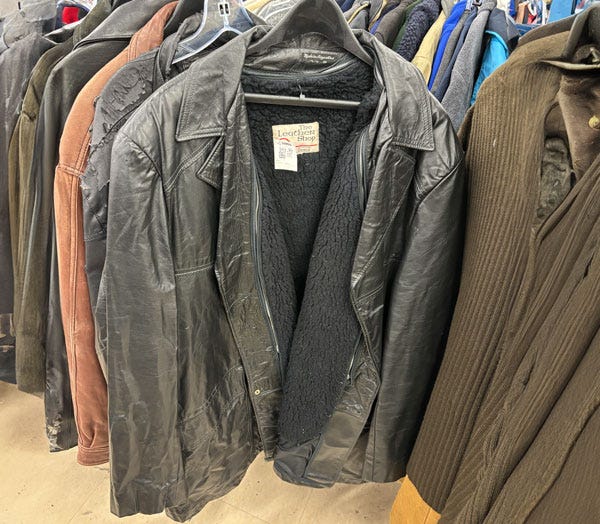 A well worn black leather jacket with a black fleece lining hangs on a thrift store rack.
