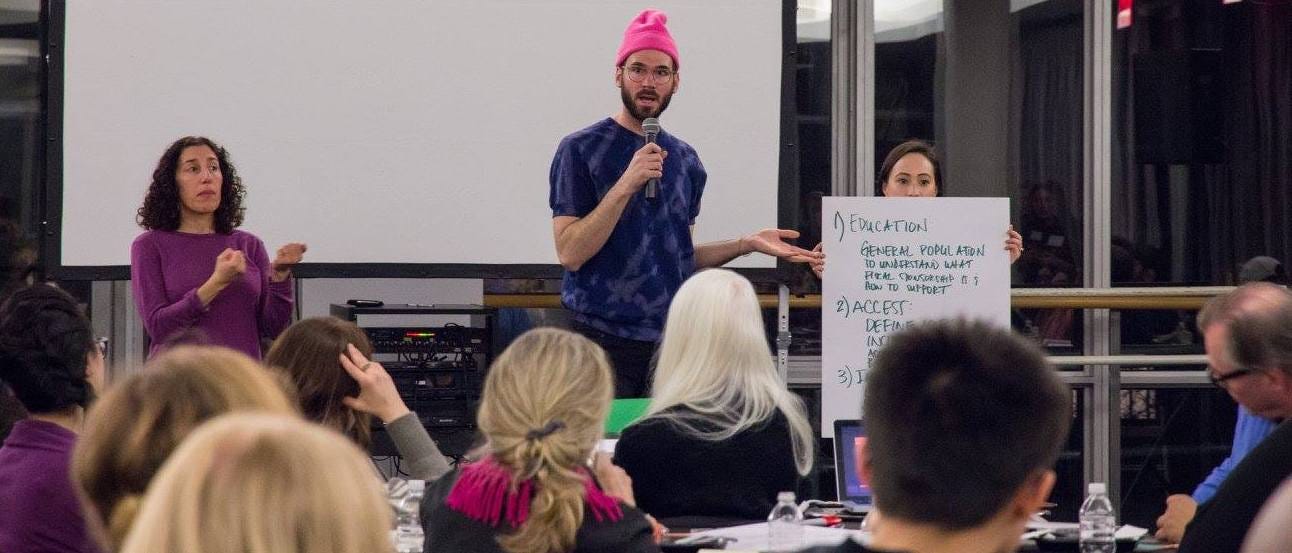 At the front of a room filled with people attending a workshop, a scruffy white person in a pink beanie hat is holding a microphone, next to a seasoned hearing ASL interpreter and a fierce white, immigrant, latina woman holding up a large piece of paper with notes, including EDUCATION and ACCESS. This speaker looks a lot like the editor of Crip News…because it’s them! Wink-face emoji.