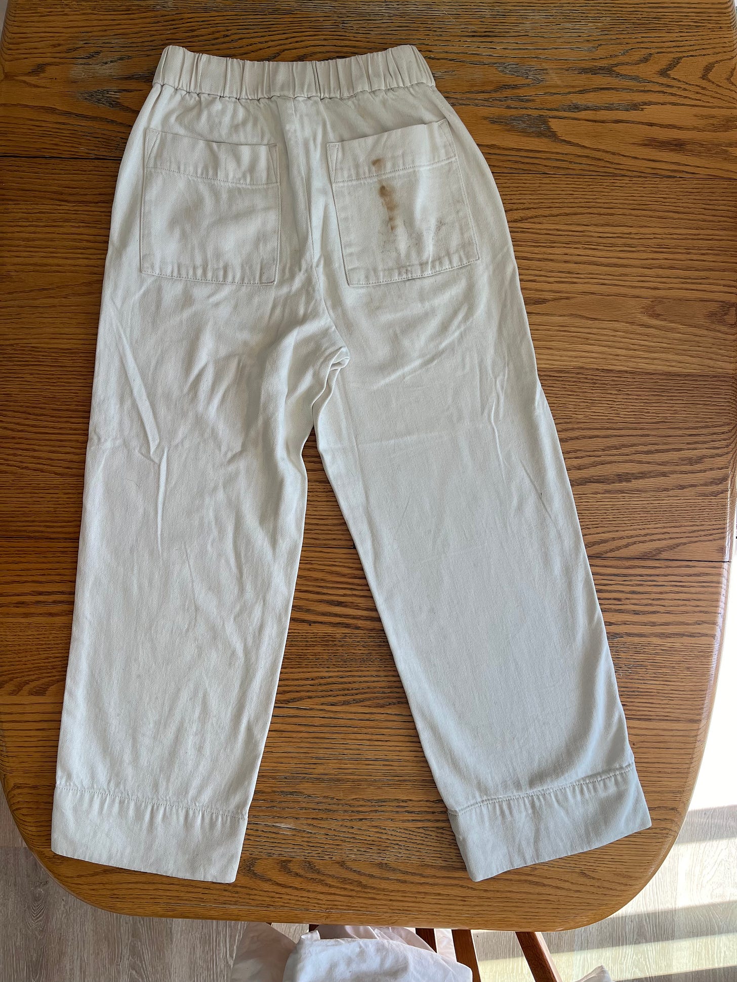 photo of a pair of white cotton pants, laying on a wooden table on a sunny day, taken from above. a small mud stain is visible on the back pocket. 