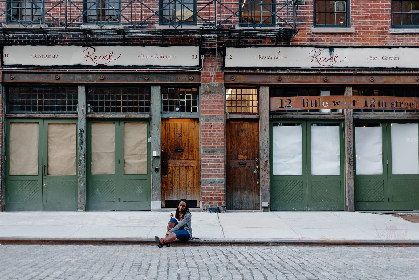  Alexa is sitting on the curb of the sidewalk holding a cup of coffee. She wears a grey sweater, jeans, and maroon boots. The buildings behind her appear to be boarded up with paper on the windows. The storefront signs say “Revel” and “little west 12th street” The doors behind her are a mix of green, and copper wood. You can also see lots of exposed brick. 