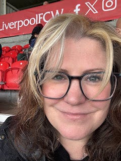 Me, in a stadium seat. The seats are red, and empty ones can be seen behind me. I've got black rimmed glasses, long brown hair with a blonde fringe, and I'm close up to the camera. Behind me head, there a red banner/sign across the back of the stand. It says Liverpool and has social media icons.