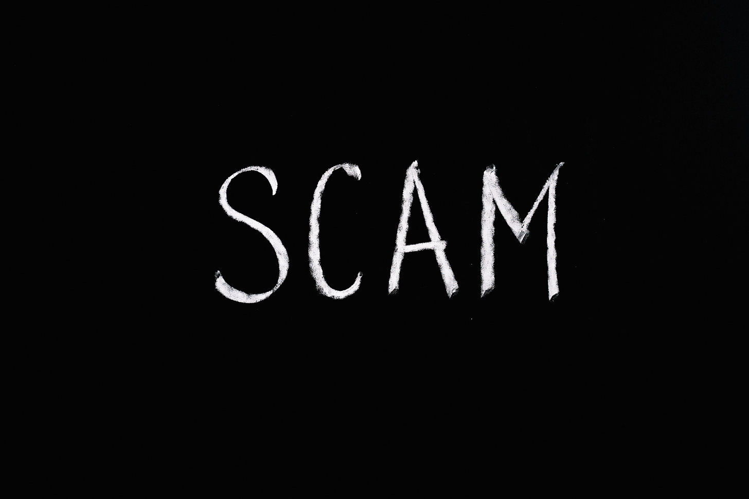 The word “SCAM” is written in white chalk on a black background.