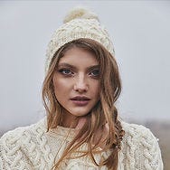 An Irish woman wearing a white Aran wool hat and jumper in front of misty mountains.